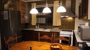 Een keuken met een houten tafel, stoelen, een koelkast, een fornuis, een spoelbak en hangende lampen.