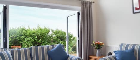 A room with two striped sofas, a small wooden table with a vase of flowers, and a view of greenery through a large window.