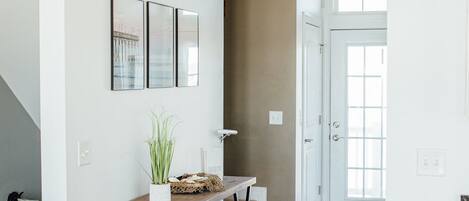 A hallway with wooden flooring, a white wall with three framed pictures, a small wooden table with a plant, and a staircase leading to another room.