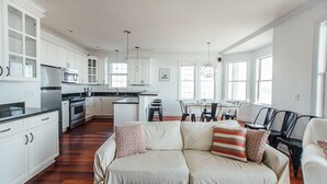 Une cuisine moderne avec des meubles blancs, un coin repas comprenant une table et des chaises, et un salon avec un canapé et des coussins.