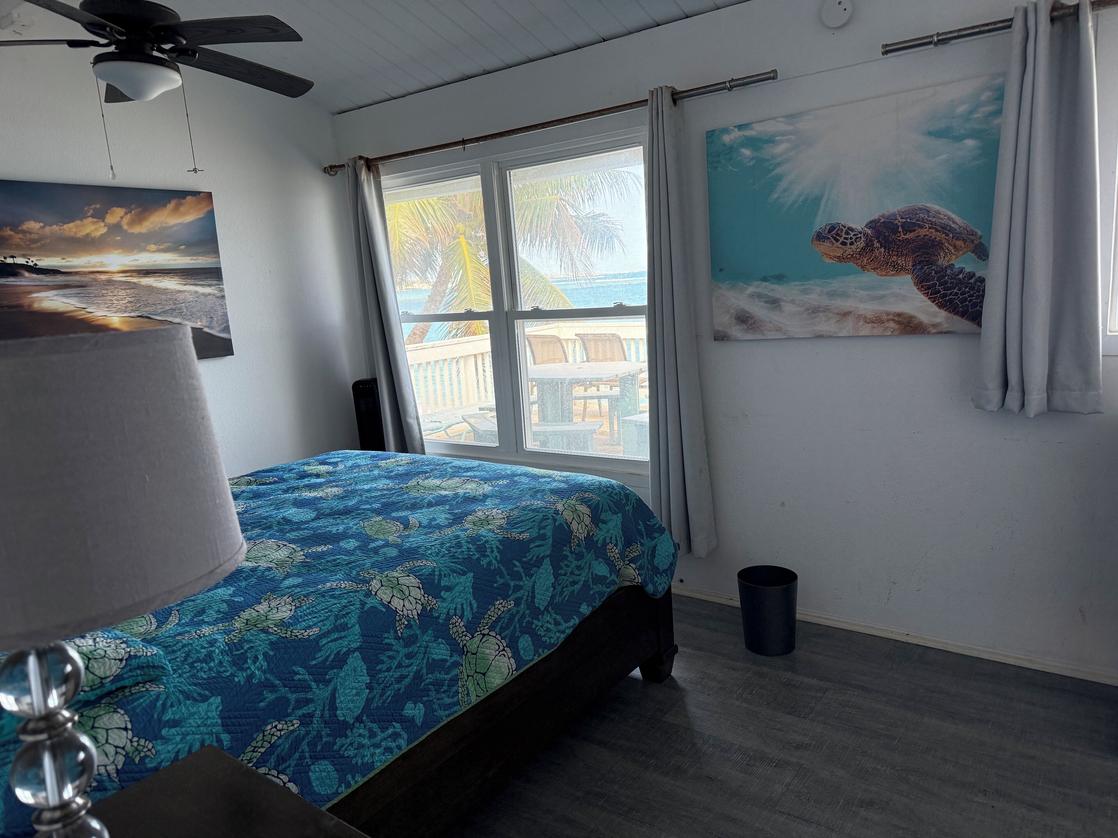 A bedroom with a bed, a ceiling fan, a window with curtains, a beach mural, and a view of the outdoors.