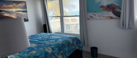 A bedroom with a bed, a ceiling fan, a window with curtains, a beach mural, and a view of the outdoors.