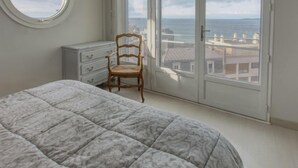 Ett sovrum med en säng, en trästol, en byrå och ett stort fönster med utsikt över byggnader och havet.