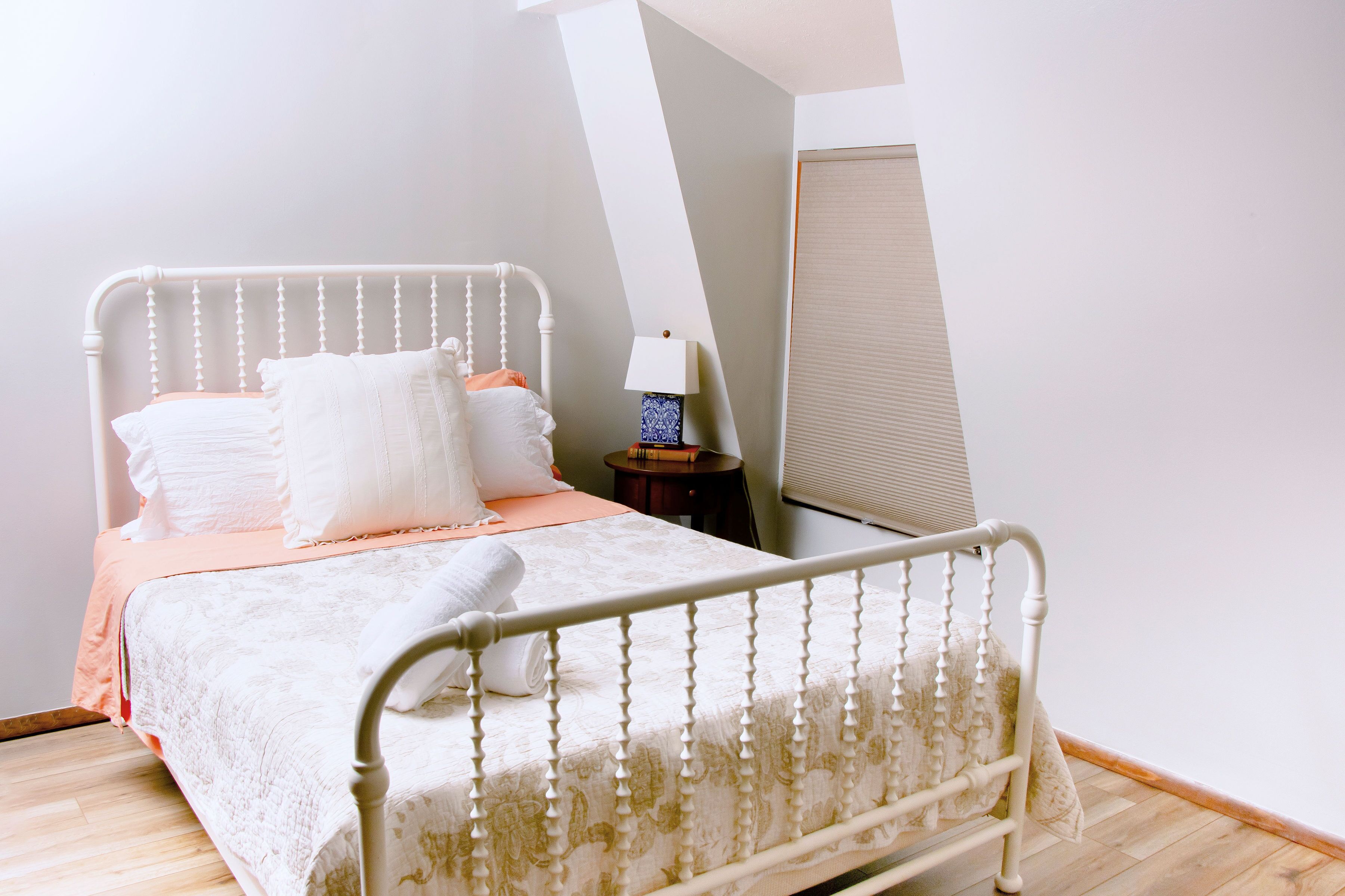 A neatly made bed with a white headboard, a bedside table with a lamp, and a window with blinds.