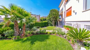 Un jardín cuidado con palmeras y diversas plantas, un césped impecable y un edificio moderno con fachada blanca y contraventanas naranjas.