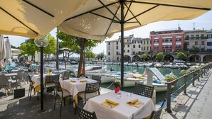 Un área de comedor al aire libre con mesas y sillas bajo sombrillas, con vista a un puerto deportivo con barcos y un hotel al fondo.