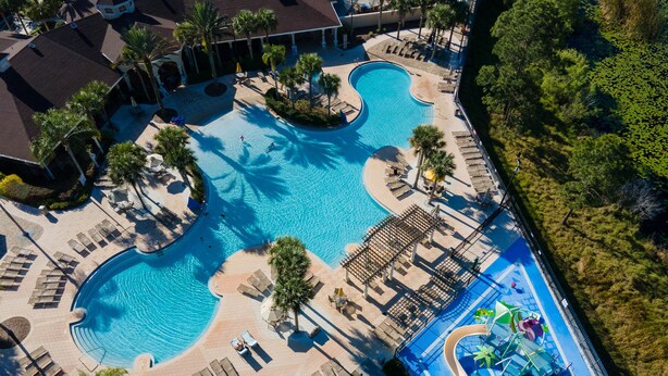 Luchtbeeld van een resort met twee grote zwembaden, ligbedden, palmbomen en een kinderspeelplaats.