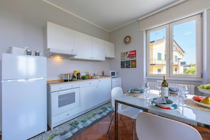 Cozinha moderna com armários brancos, sala de jantar com mesa posta para dois e janela com vista para edifícios.