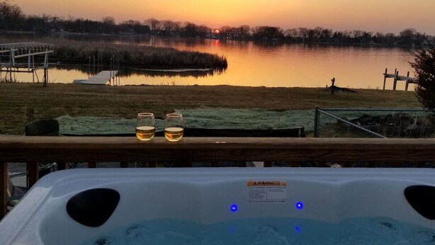 A hot tub with two glasses of beer on a wooden deck overlooking a lake at sunset.