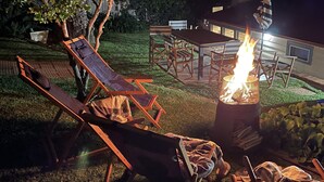 Uma área de estar ao ar livre com espreguiçadeiras e uma mesa, um poço de fogo e uma casa ao fundo.