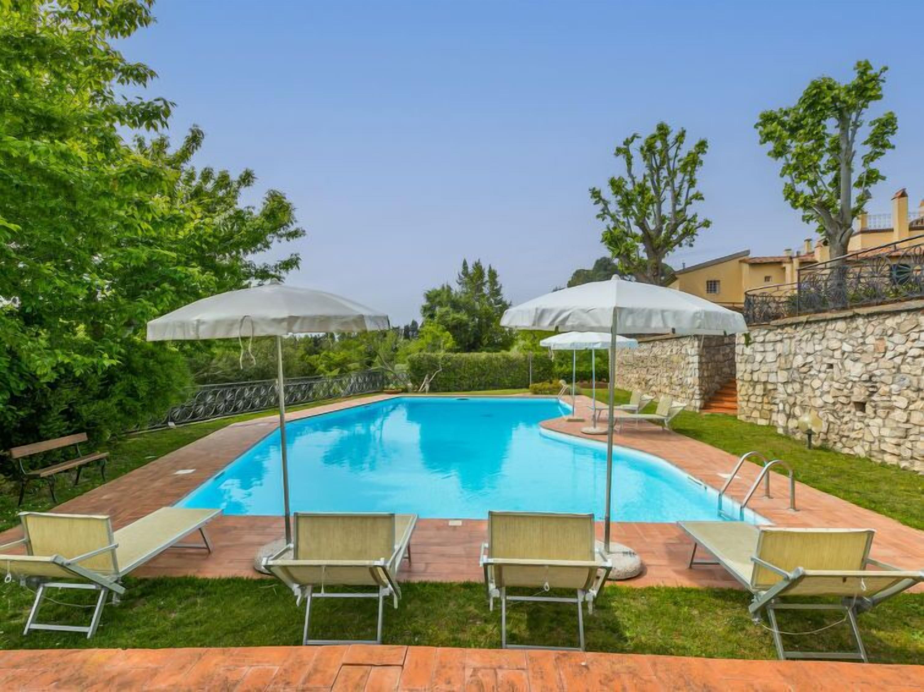 Una piscina rectangular con un muro de piedra y una zona de césped con sillones y sombrillas.