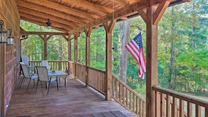 A covered wooden deck with a table and chairs, surrounded by trees and an American flag. - Peaceful Tignall Cabin on Strom Thurmond Lake! (Tignall)