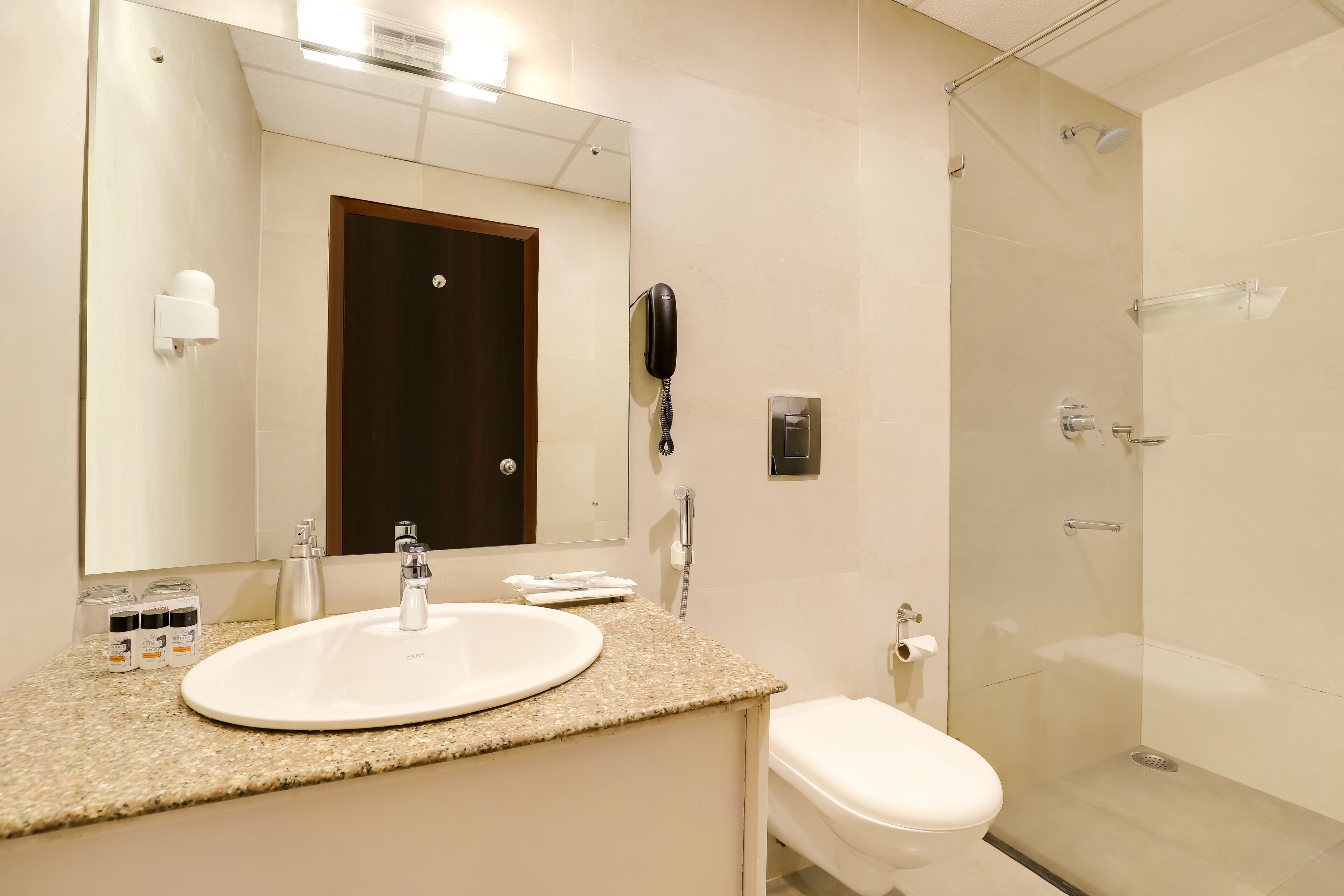 A modern bathroom with a white sink, a toilet, a glass-enclosed shower, and a mirror with a phone mounted on the wall.