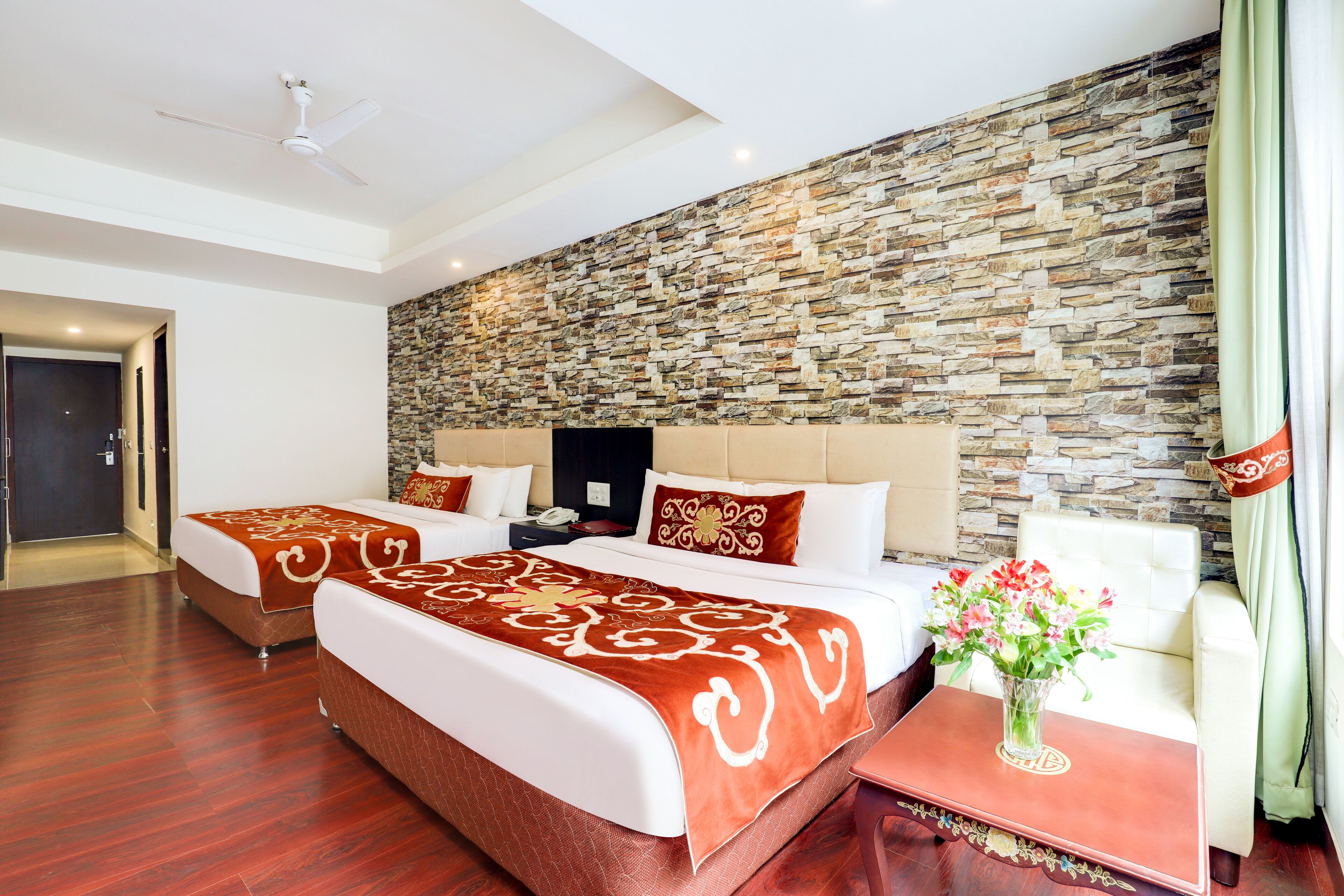 A hotel room with a stone wall, two beds with red and white bedding, a wooden floor, a white ceiling fan, a nightstand with a vase of flowers, and a green curtain.