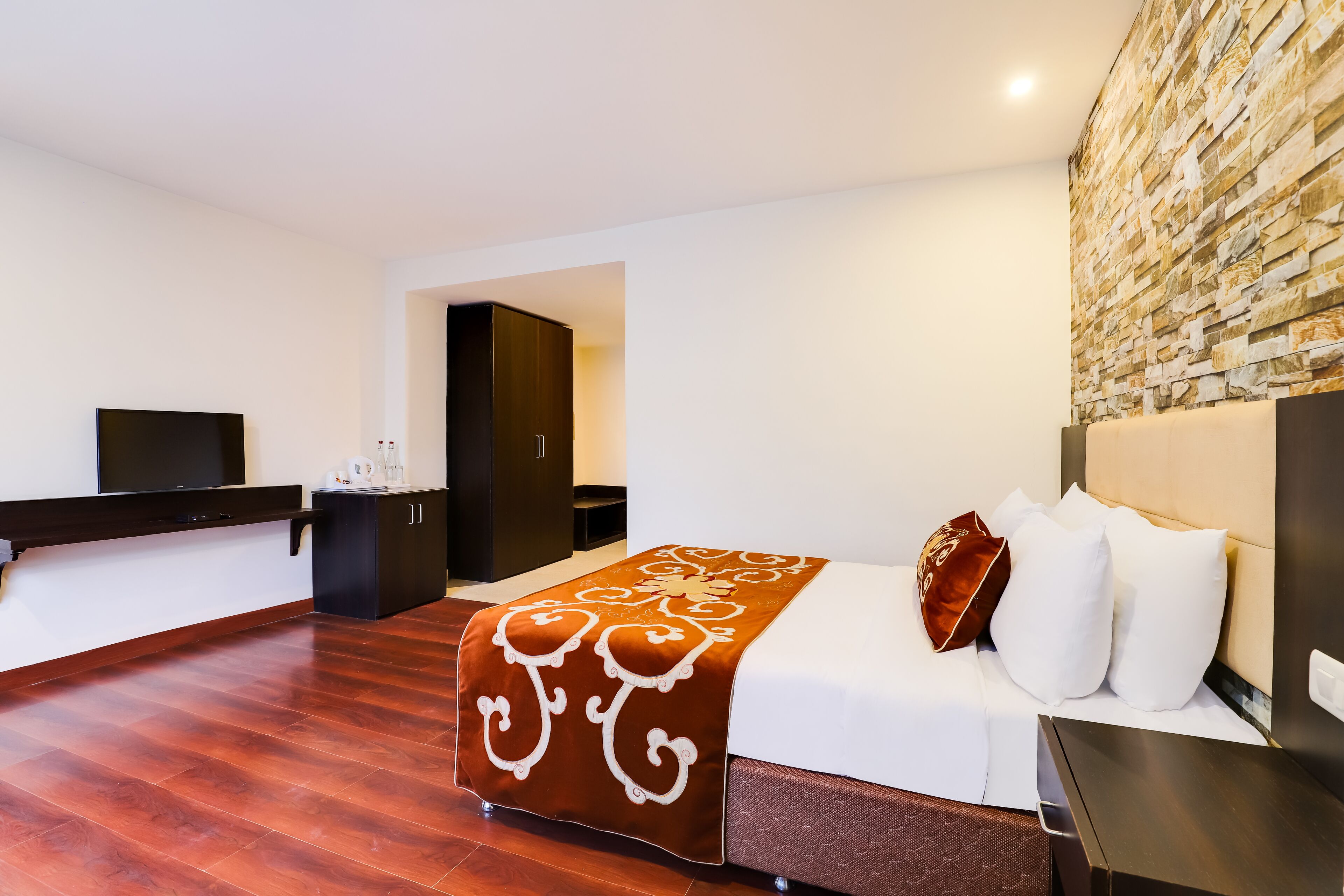 A modern hotel room with a wooden floor, a large bed with white and brown bedding, a flat-screen TV, a dark wooden desk, and a stone-textured wall.