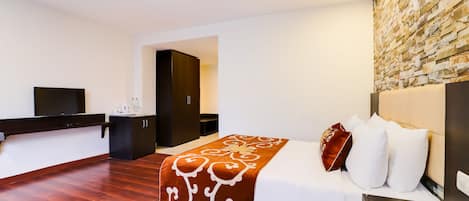 A modern hotel room with a wooden floor, a large bed with white and brown bedding, a flat-screen TV, a dark wooden desk, and a stone-textured wall.