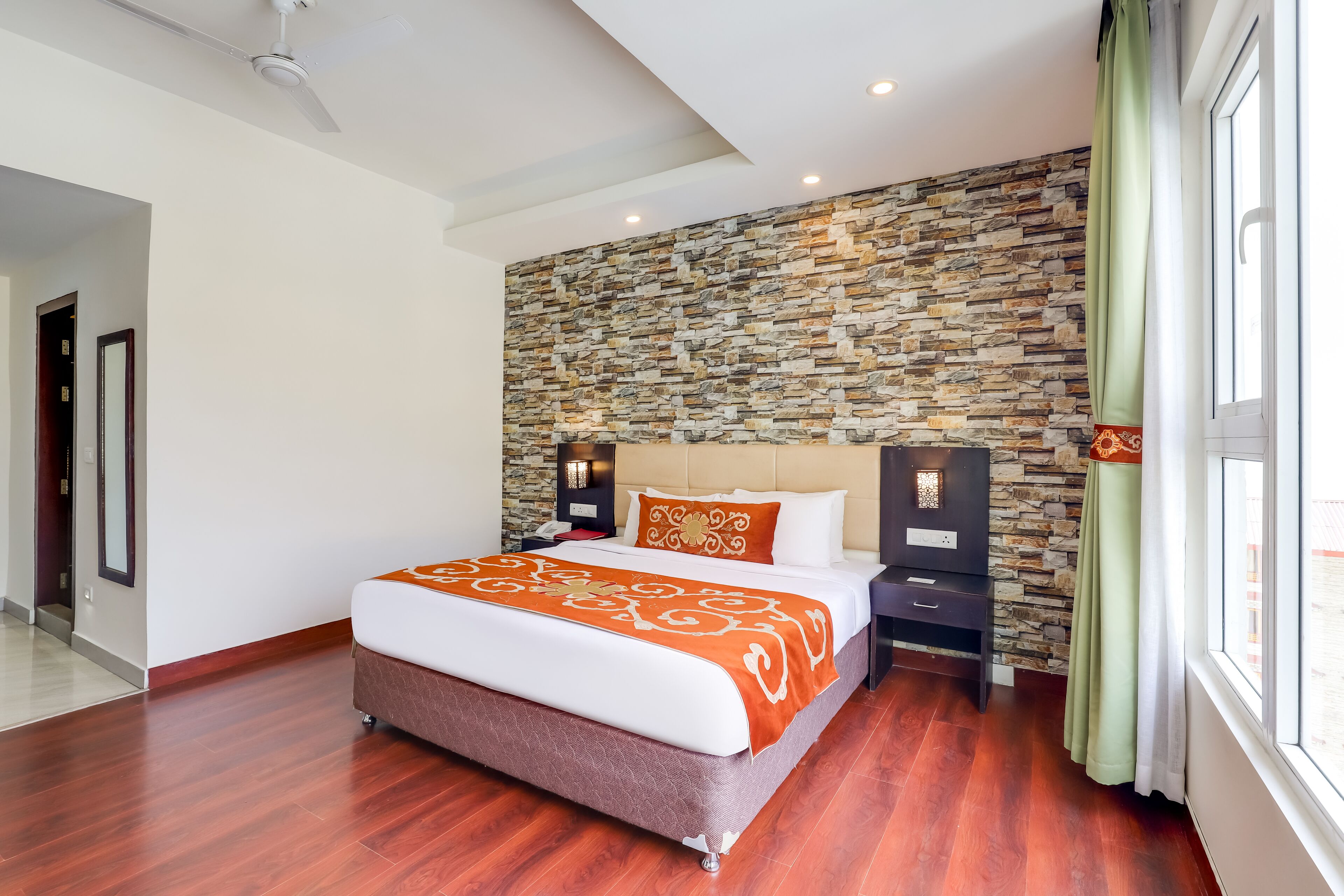 A modern hotel room with a large bed, stone wall, wooden flooring, and a view of the city.