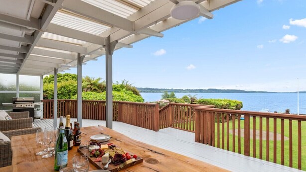 A wooden deck with a table set for dining, overlooking a body of water with a view of the horizon. - Wake Up by the Lake | Private Jetty, Kayaks & Cosy Comforts (Rotorua)