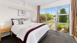 A bedroom with a large bed, a nightstand, a window with a view of a garden, and a painting on the wall. - Wake Up by the Lake | Private Jetty, Kayaks & Cosy Comforts (Rotorua)
