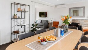 A modern living room with a dining area, featuring a sofa, a coffee table with breakfast items, and a black metal shelf with decorative items. - Wake Up by the Lake | Private Jetty, Kayaks & Cosy Comforts (Rotorua)