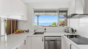 A modern kitchen with white cabinets, a stainless steel dishwasher, a built-in oven, and a window with a view of the sea. - Wake Up by the Lake | Private Jetty, Kayaks & Cosy Comforts (Rotorua)