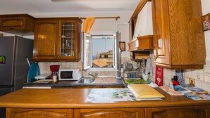 A kitchen with wooden cabinets, a refrigerator, a microwave, and a window with a view of buildings. - City walls sea view (Corfu)