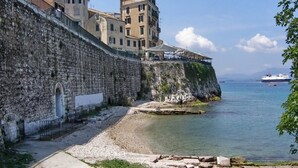 A coastal stone wall with buildings on top, overlooking a calm sea with a ship and a small boat. - City walls sea view (Corfu)