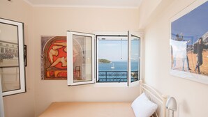 A bedroom with a bed, a nightstand, a lamp, a window with an open view of the sea, and a painting on the wall. - City walls sea view (Corfu)