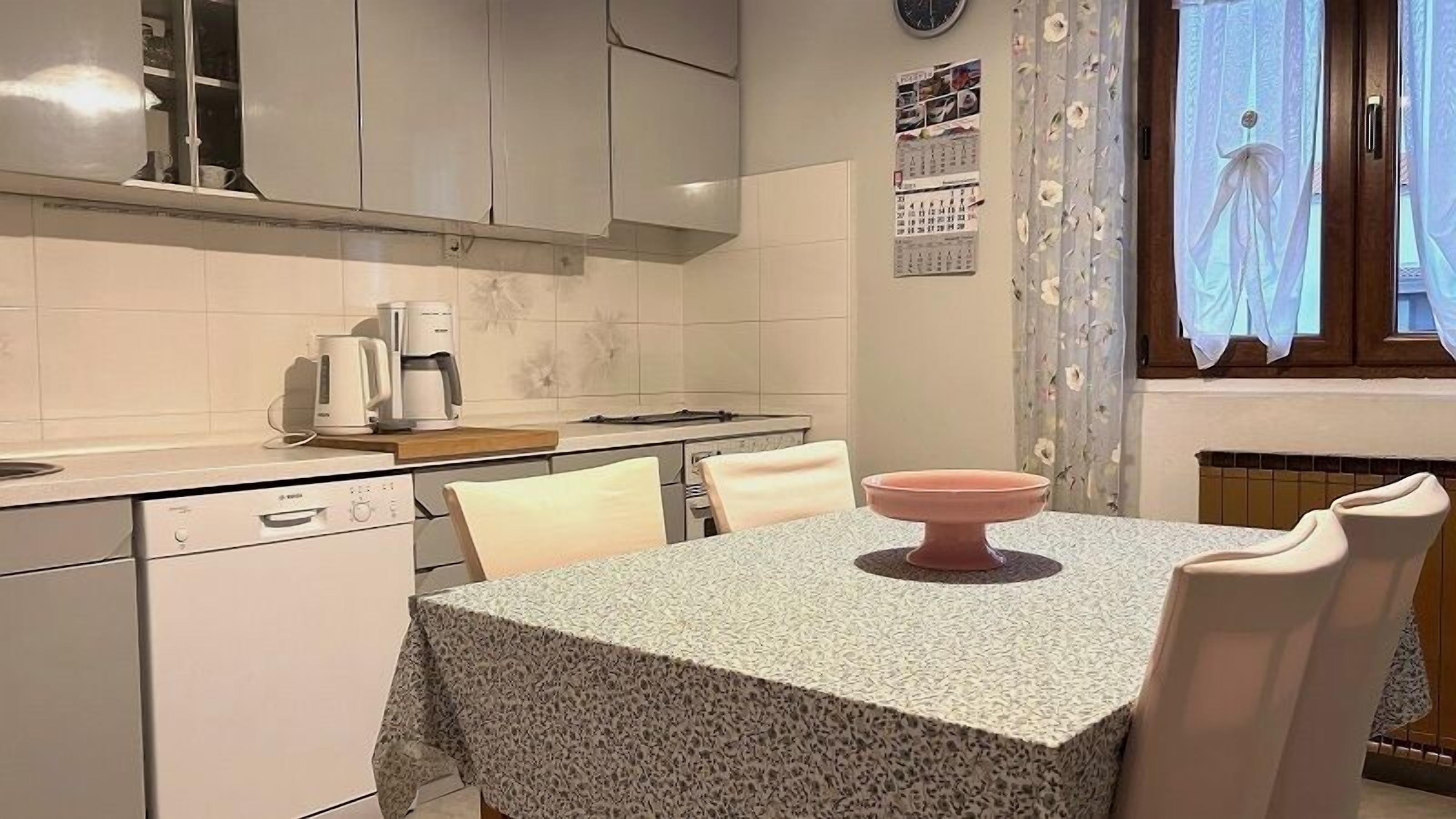 A compact kitchen with white cabinets, a coffee maker, a calendar, and a window with floral curtains.