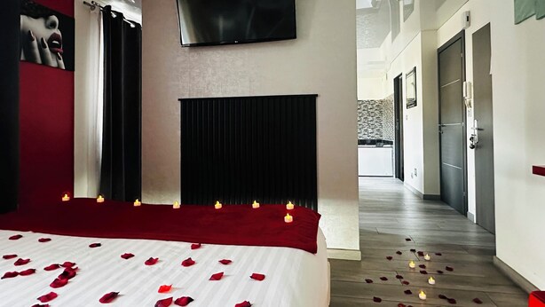 Una habitación de hotel con una cama, un televisor montado en la pared, una alfombra roja y un pasillo que da a otras habitaciones.