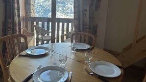 Ein Esstisch mit vier Stühlen, gedeckt für vier Personen, in einem Zimmer mit Blick auf eine verschneite Berglandschaft durch ein Fenster mit gemusterten Vorhängen.