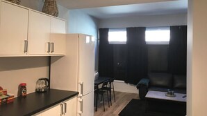 A compact kitchen with white cabinets, a black countertop, and a refrigerator. There is a dining area with a table and chairs, and a black sofa. - Notodden Sentrum Apartment NO 10 (Notodden)