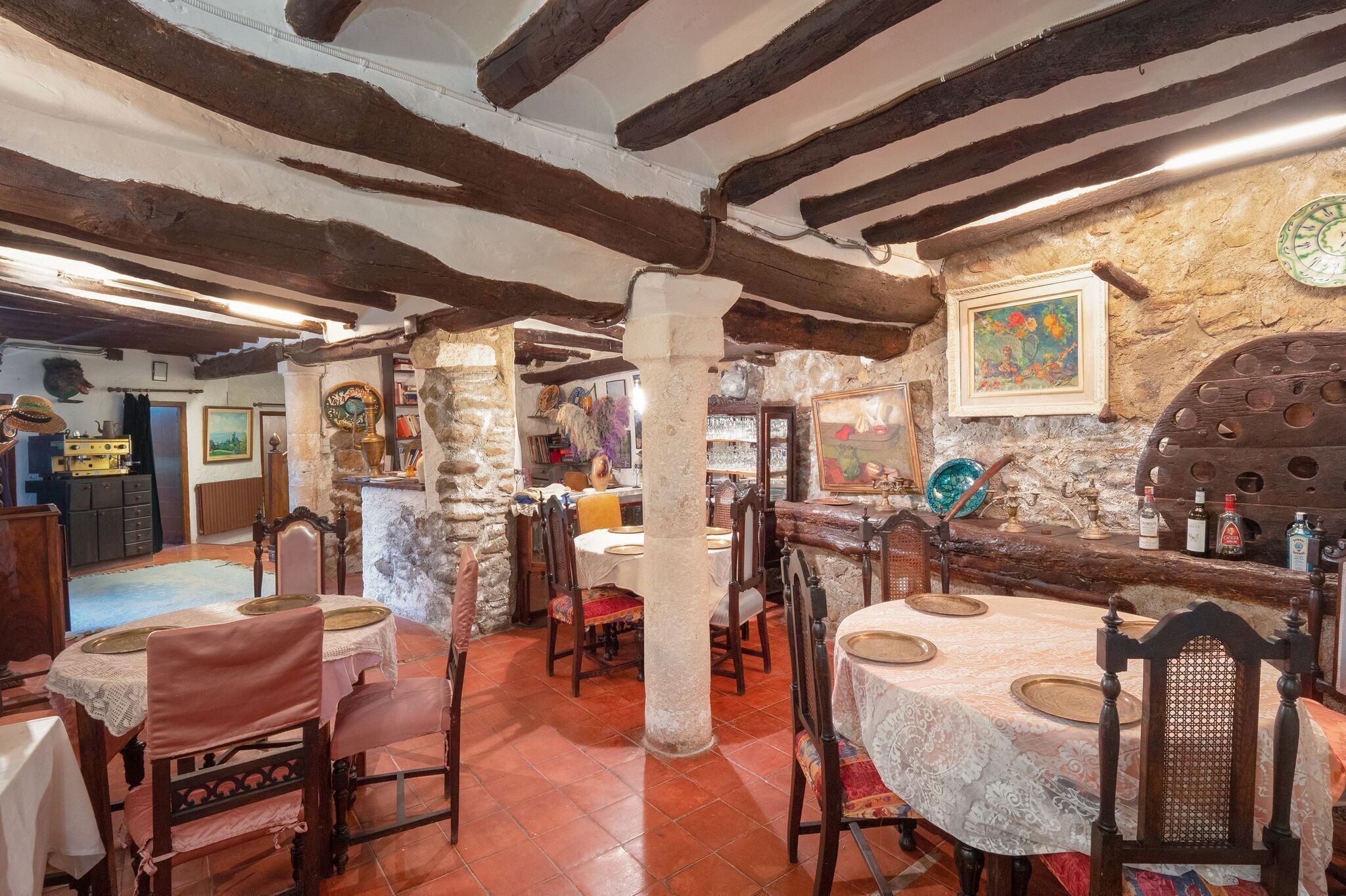 Un comedor rústico con vigas de madera a la vista, paredes de piedra y decoración tradicional. Las mesas están dispuestas con manteles blancos y platos de madera.