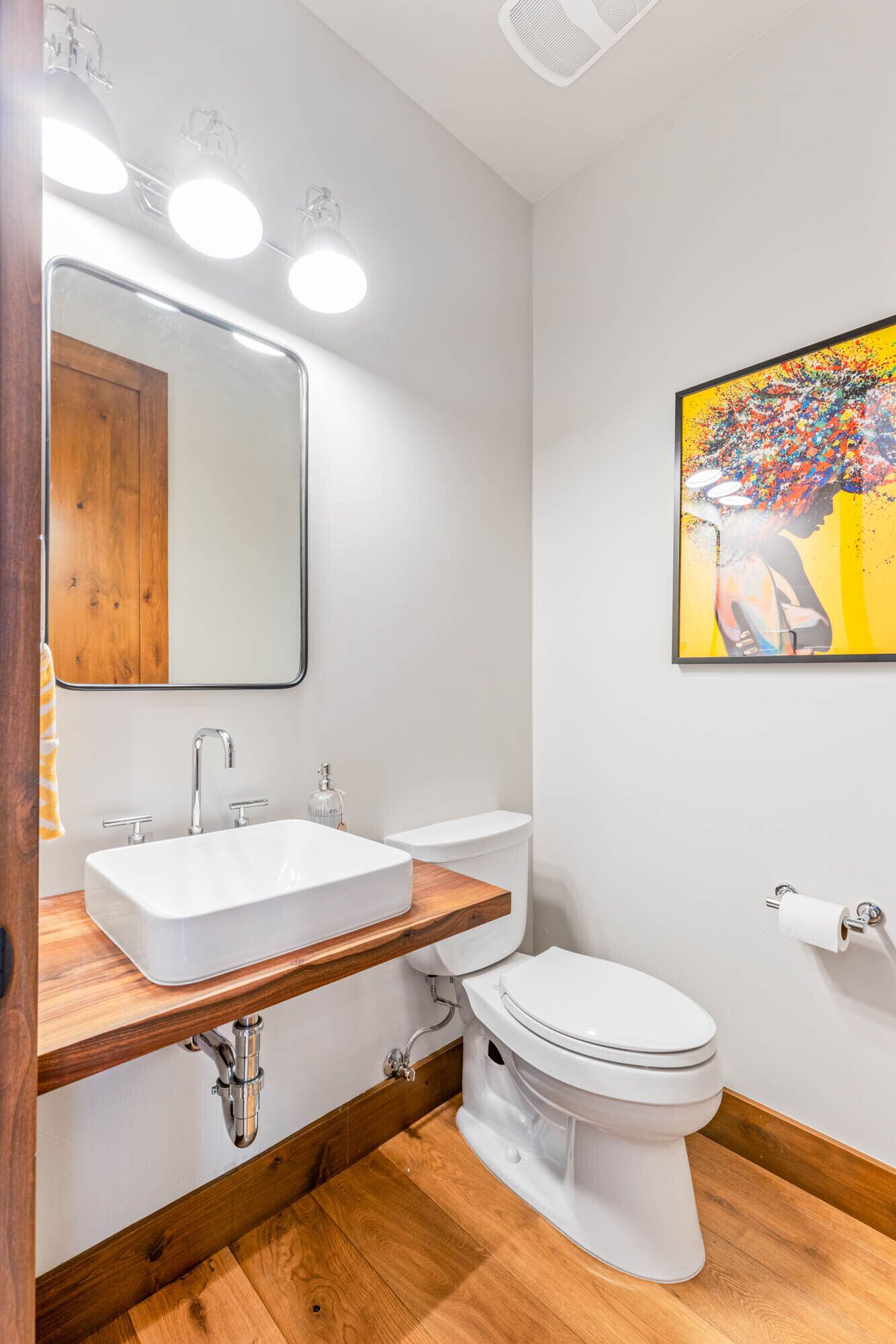A modern bathroom with a white sink, a mirror, and a toilet. There is a wooden vanity and a painting on the wall.