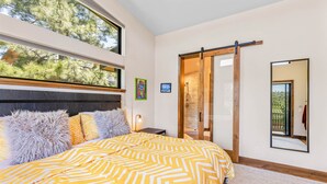 A modern bedroom with a large bed, a sliding glass door to a balcony, and a view of trees.