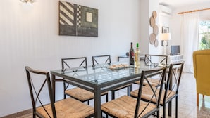 Een eethoek met een glazen tafel, geweven stoelen, een schilderij aan de muur, een lamp en een televisie.