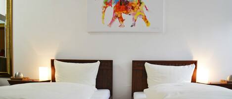 A hotel room with two beds, white linens, wooden headboards, a wall-mounted painting of a colorful elephant, and bedside lamps.