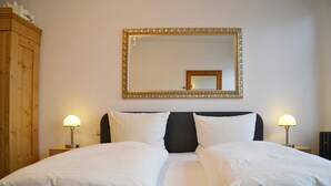 Uma cama bem arrumada com lençóis brancos, duas luminárias de cabeceira, um espelho grande com moldura ornamentada e um guarda-roupa de madeira.