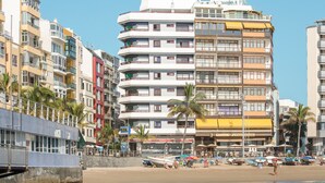Ein mehrstöckiges Hotel mit Balkonen, ein Strand im Vordergrund und Palmen.
