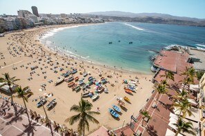 A beach with rows of umbrellas and boats, a promenade with palm trees, and a cityscape in the background.