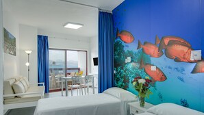 Una habitación de hotel con dos camas, una zona de comedor con mesa y sillas, vista al mar y un mural de peces en la pared.