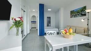 Una habitación de hotel moderna con zona de comedor, televisor, un cuadro y un frutero.