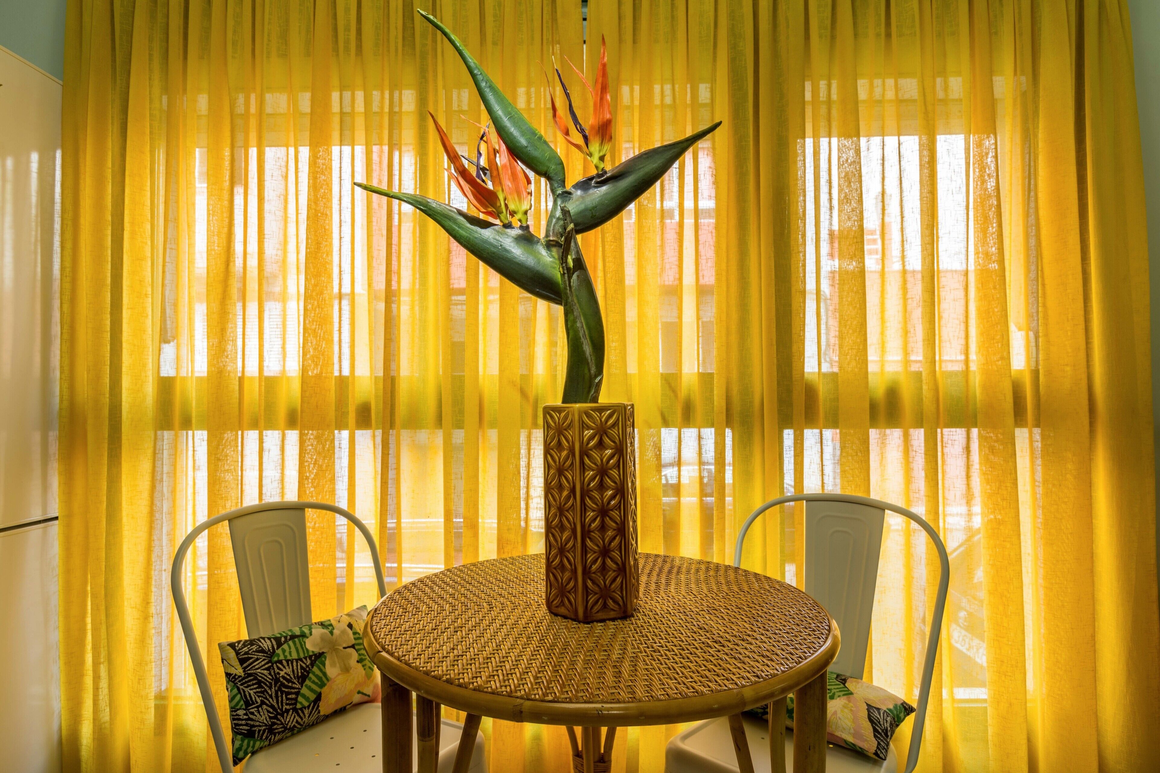 Una mesa redonda con superficie tejida, dos sillas blancas, un florero decorativo con planta y cortinas amarillas al fondo.