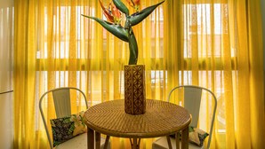 A round table with a woven surface, two white chairs, a decorative vase with a plant, and yellow curtains in the background.