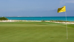 Campo de golf con bandera, vista al océano y playa arenosa.