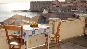 A table with a checkered tablecloth, two wooden chairs, and a basket of fruit on a terrace overlooking a waterfront with a historic building in the background. - Room Lazareti (Dubrovnik)