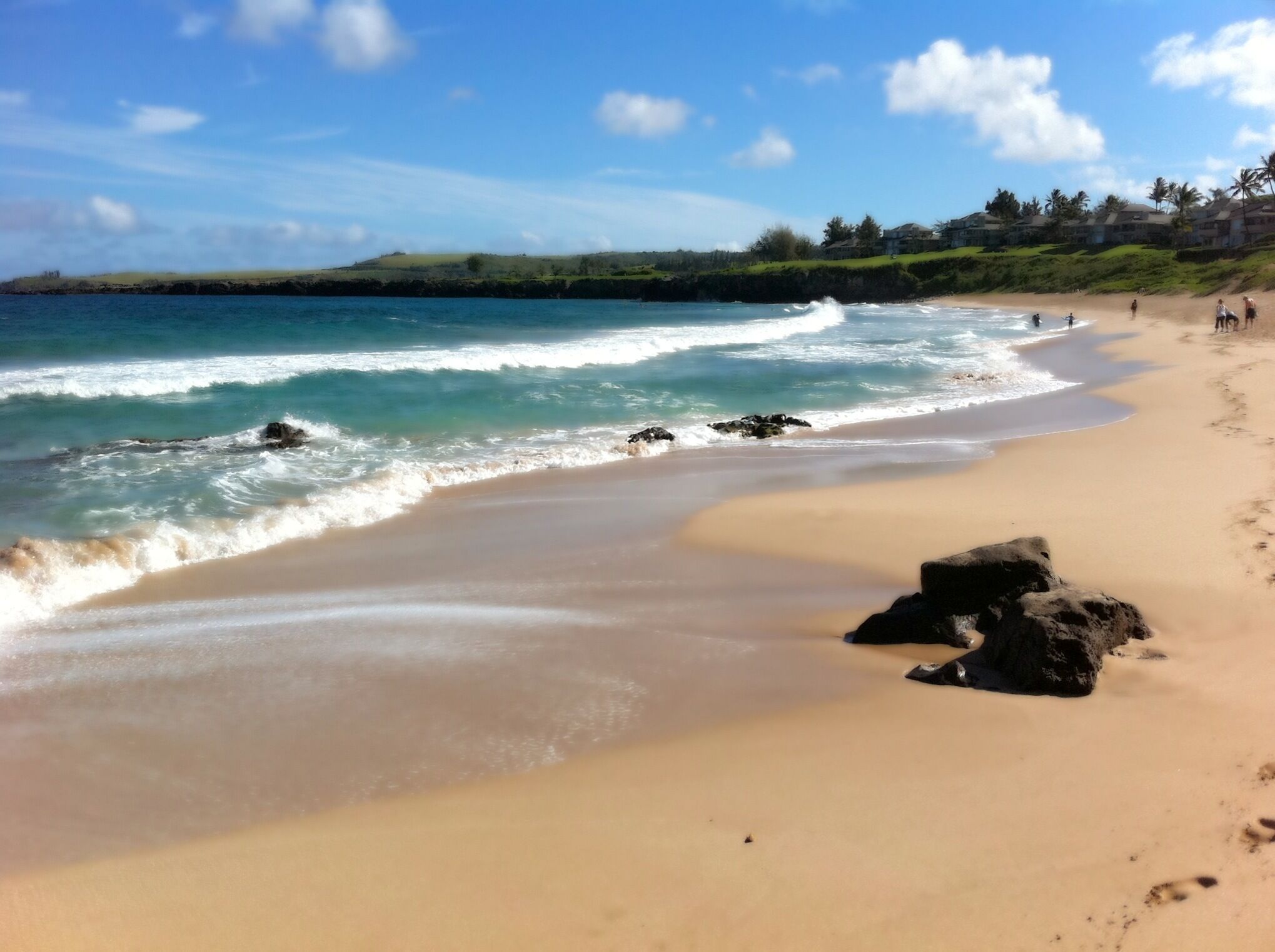 A beach with clear blue water, sandy shore, and a few people walking.
