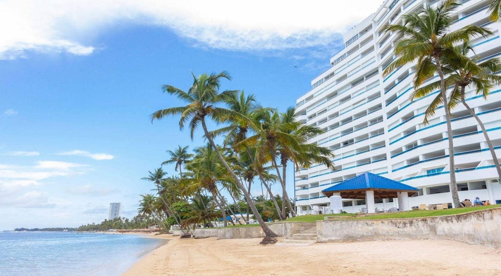 Un hotel frente a la playa con una estructura de techo azul, palmeras y una playa de arena.