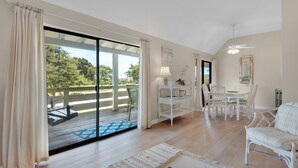 Ein geräumiges Wohnzimmer mit einem großen Schiebeglasfenster, das auf eine Terrasse führt, einem Essbereich mit weißer Möbel und einem Deckenventilator.