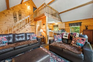 A cozy cabin interior with a wooden staircase, leather sofas, and a kitchen area. - Lupine Lodge - Adorable, newly renovated cottage, with water access and dock on Loon Lake (Rangeley)
